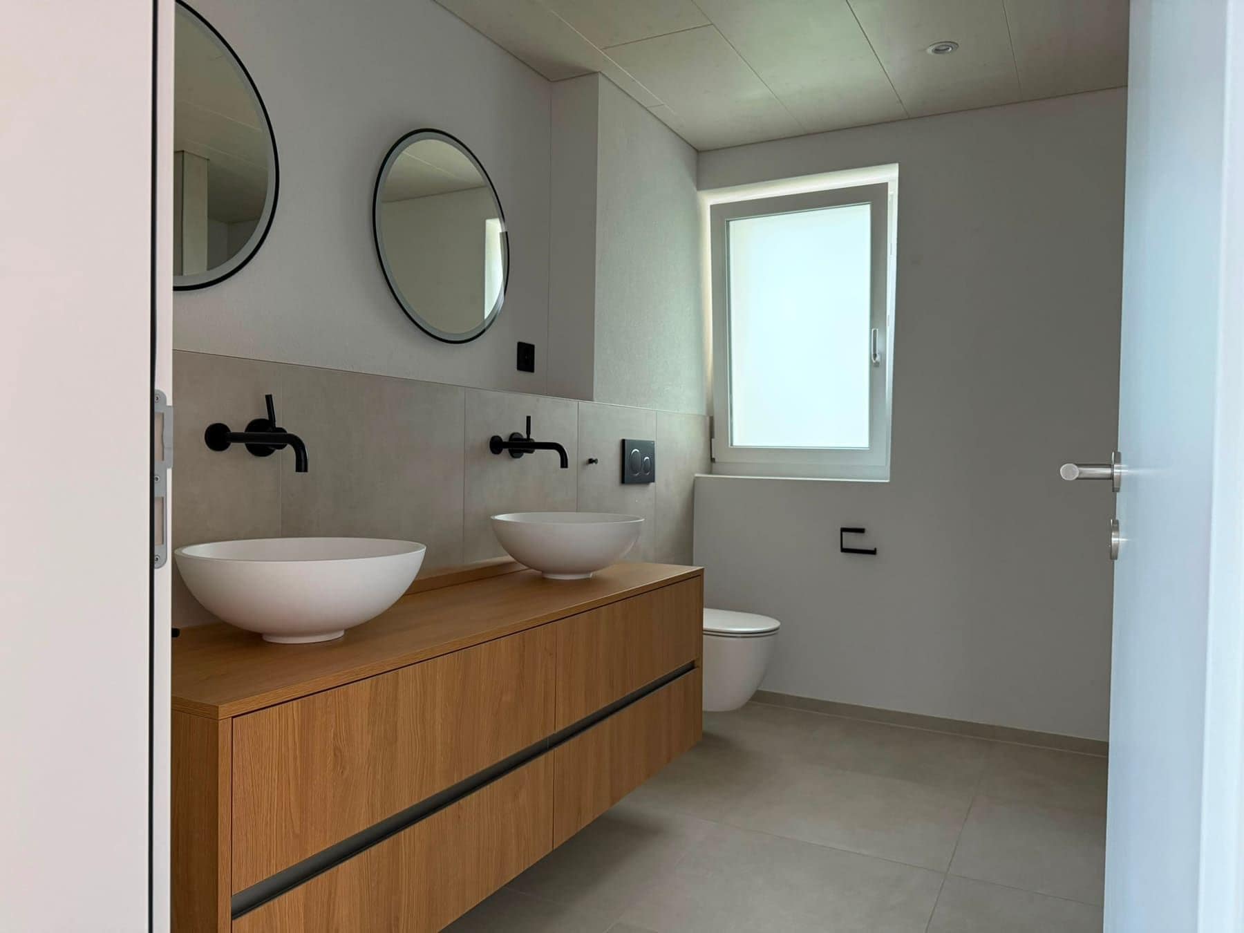 Neues Badezimmer mit zwei Aufsatzbecken auf einem Holzmöbel, runden Spiegeln und Fenster
