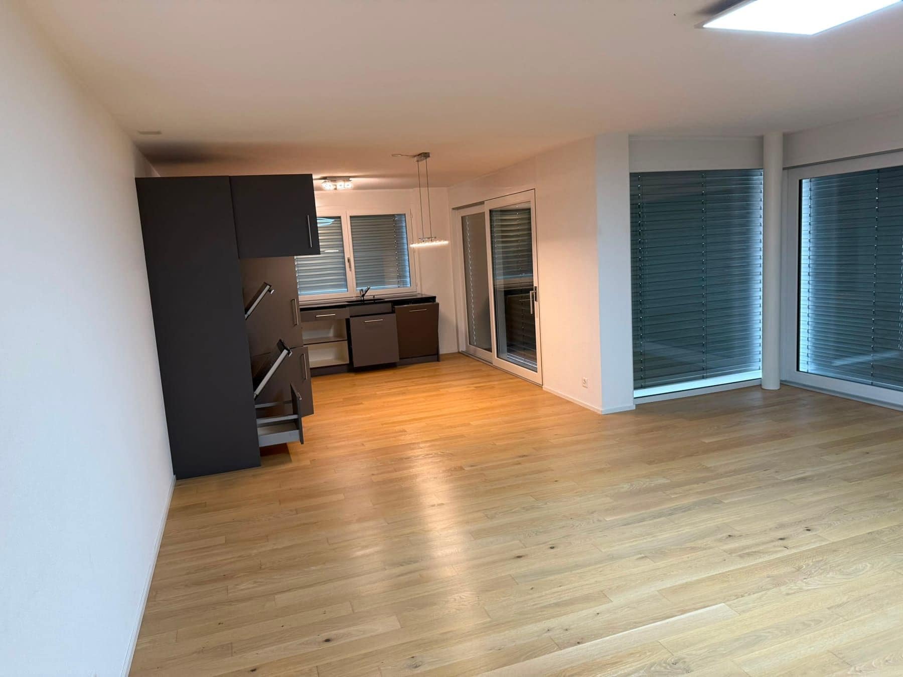 Leere Wohnung mit Parkett, Küchenzeile und Fensterfront mit Lamellenstoren